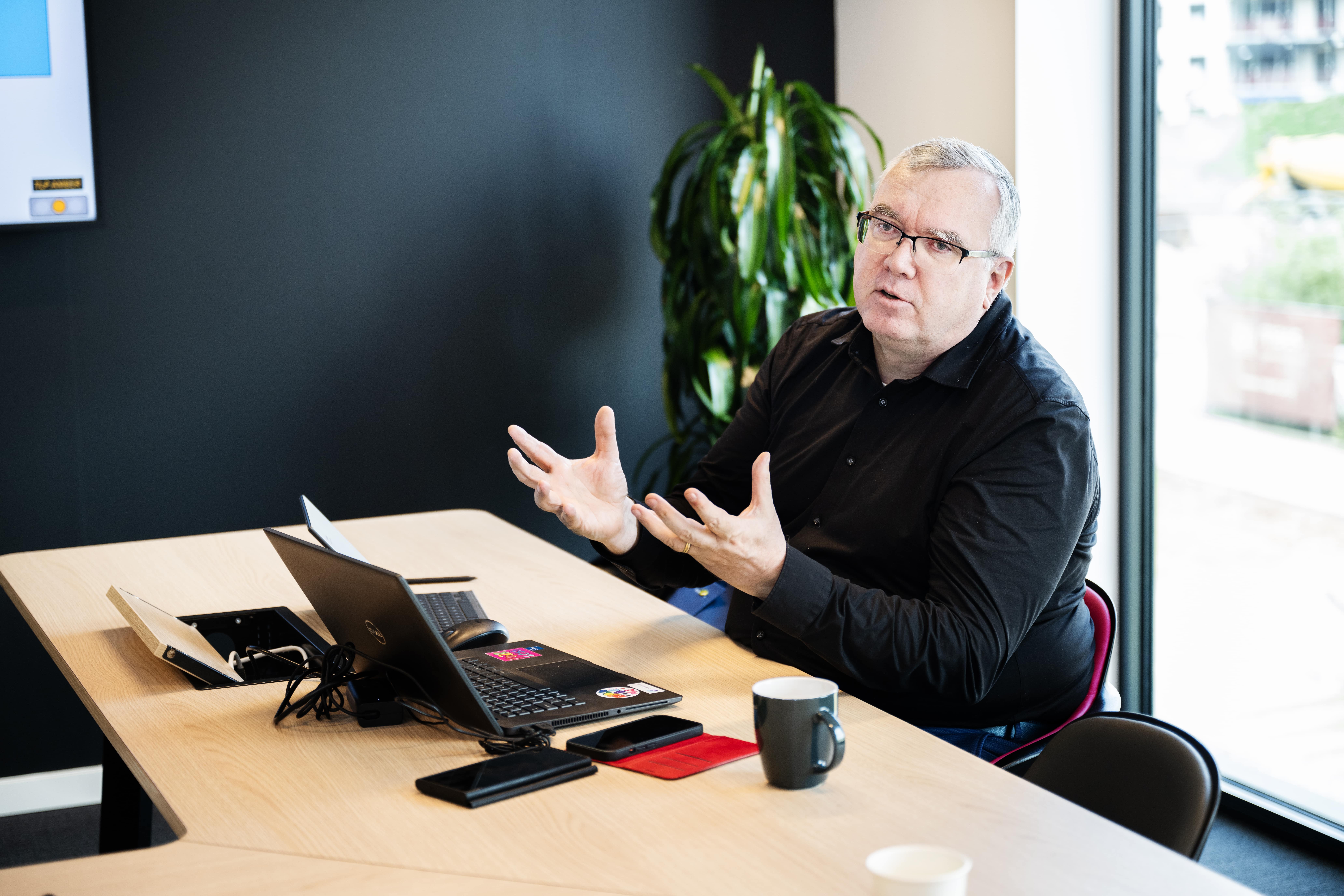 Man gesturing while speaking in modern office setting, seated at a conference table with a laptop and coffee mug, large window and plant visible in background