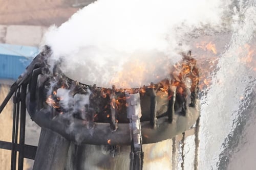 Industrial chimney with fire at the edge, emitting white smoke against a blurred background