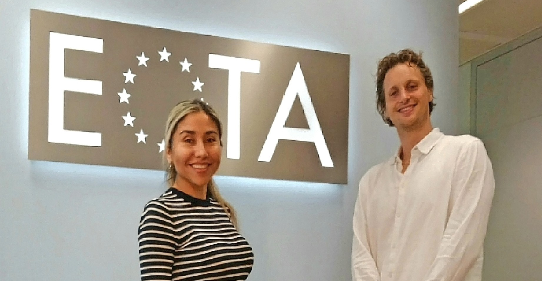 Two people smiling in front of an EOTA sign in an office setting, symbolizing teamwork and collaboration