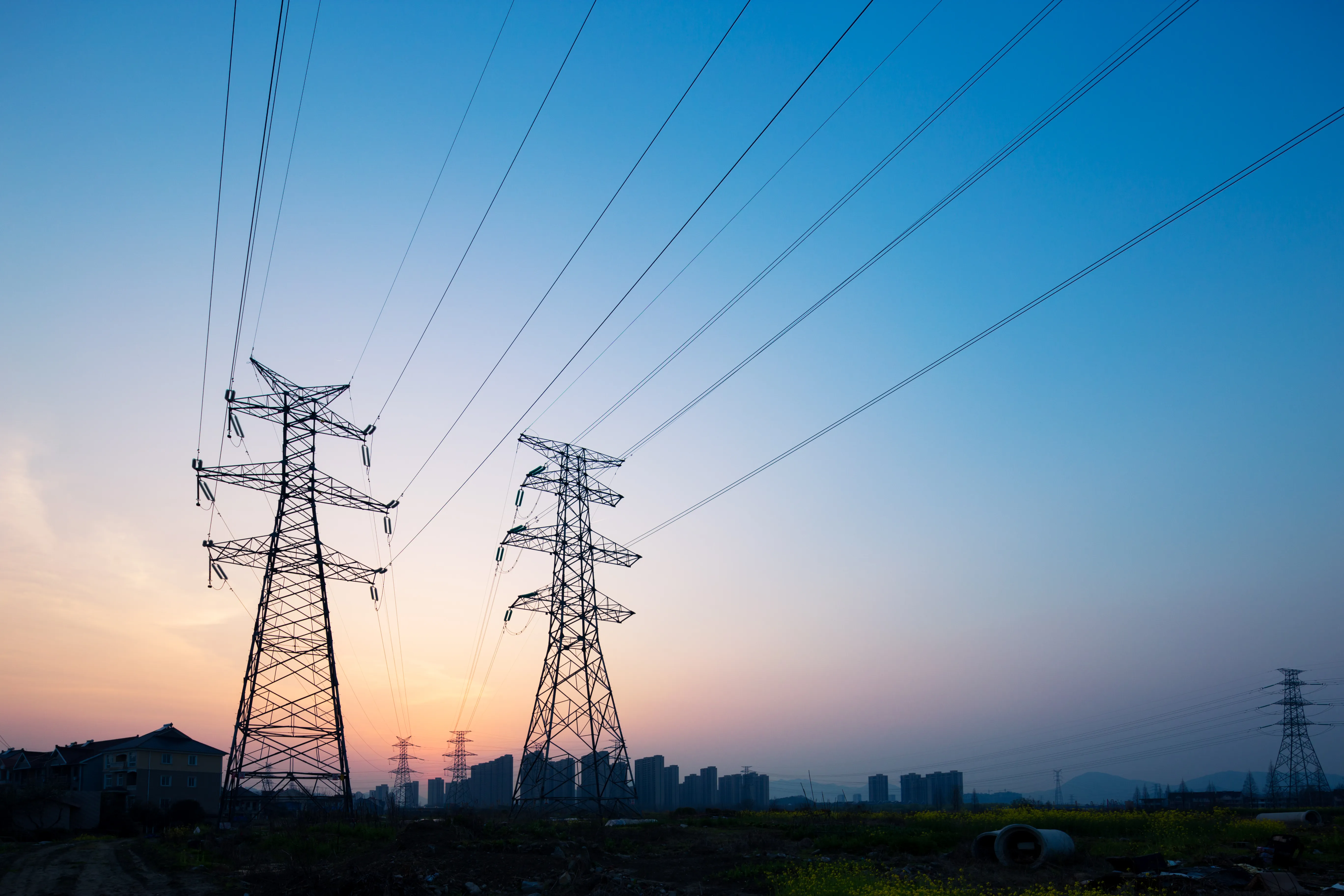 High-voltage power lines and transmission towers at sunset, silhouetted against a colorful sky, with a city skyline in the background