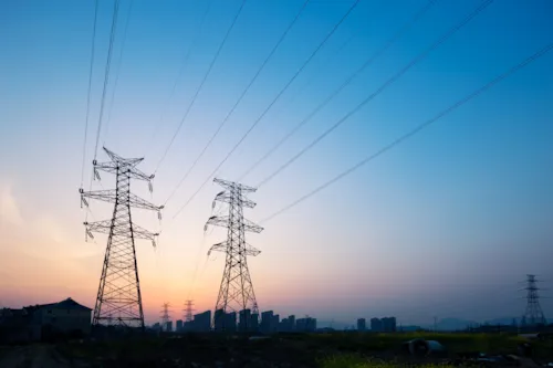 High-voltage power lines and transmission towers at sunset, silhouetted against a colorful sky, with a city skyline in the background