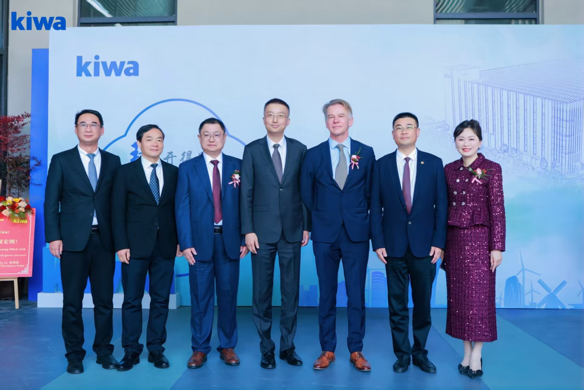 Seven people in formal attire stand in a row at a Kiwa event, set against a backdrop featuring the Kiwa logo and architectural illustrations