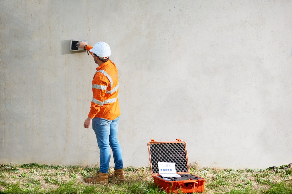 Kiwa inspector using a handheld device to inspect a concrete wall, wearing an orange safety jacket and white hard hat, with an open equipment case nearby