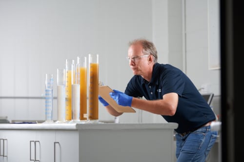 Kiwa laboratory employee examining test tubes with liquid samples in a laboratory, wearing safety goggles and gloves, holding a clipboard for notes