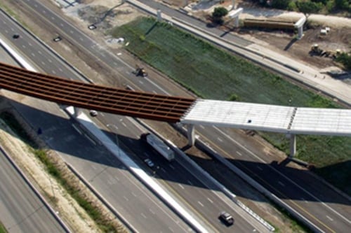 Aerial view of an unfinished overpass bridge under construction spanning a multi-lane highway, with vehicles passing below and surrounding green spaces