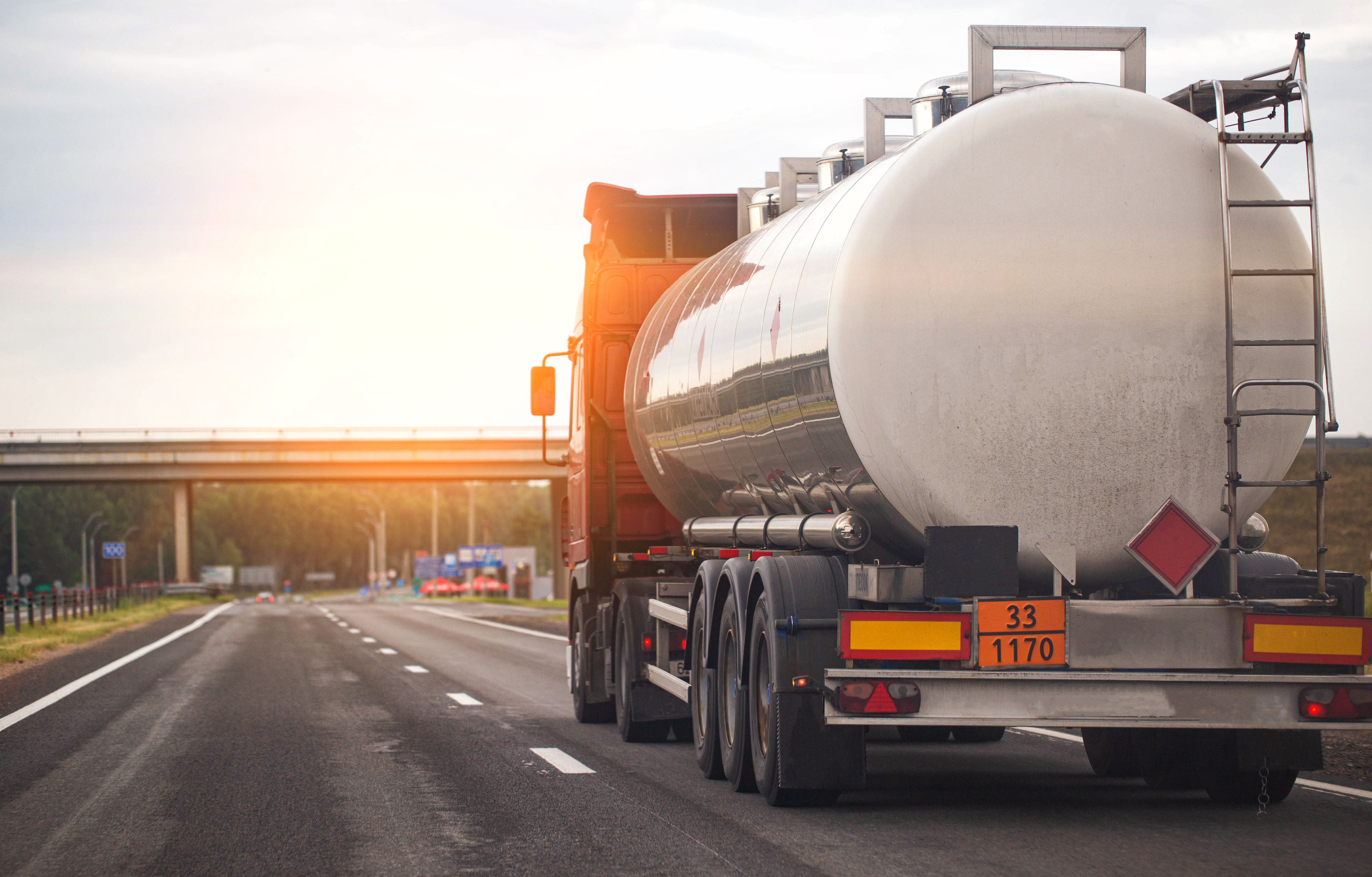 Tanker truck transporting liquid cargo on a highway during sunset, with a bridge in the background