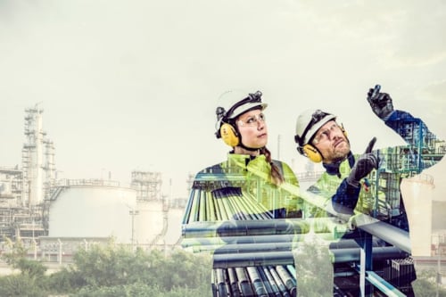 Industrial engineers wearing safety gear and helmets, examining processes at a petrochemical plant with industrial equipment and storage tanks in the background