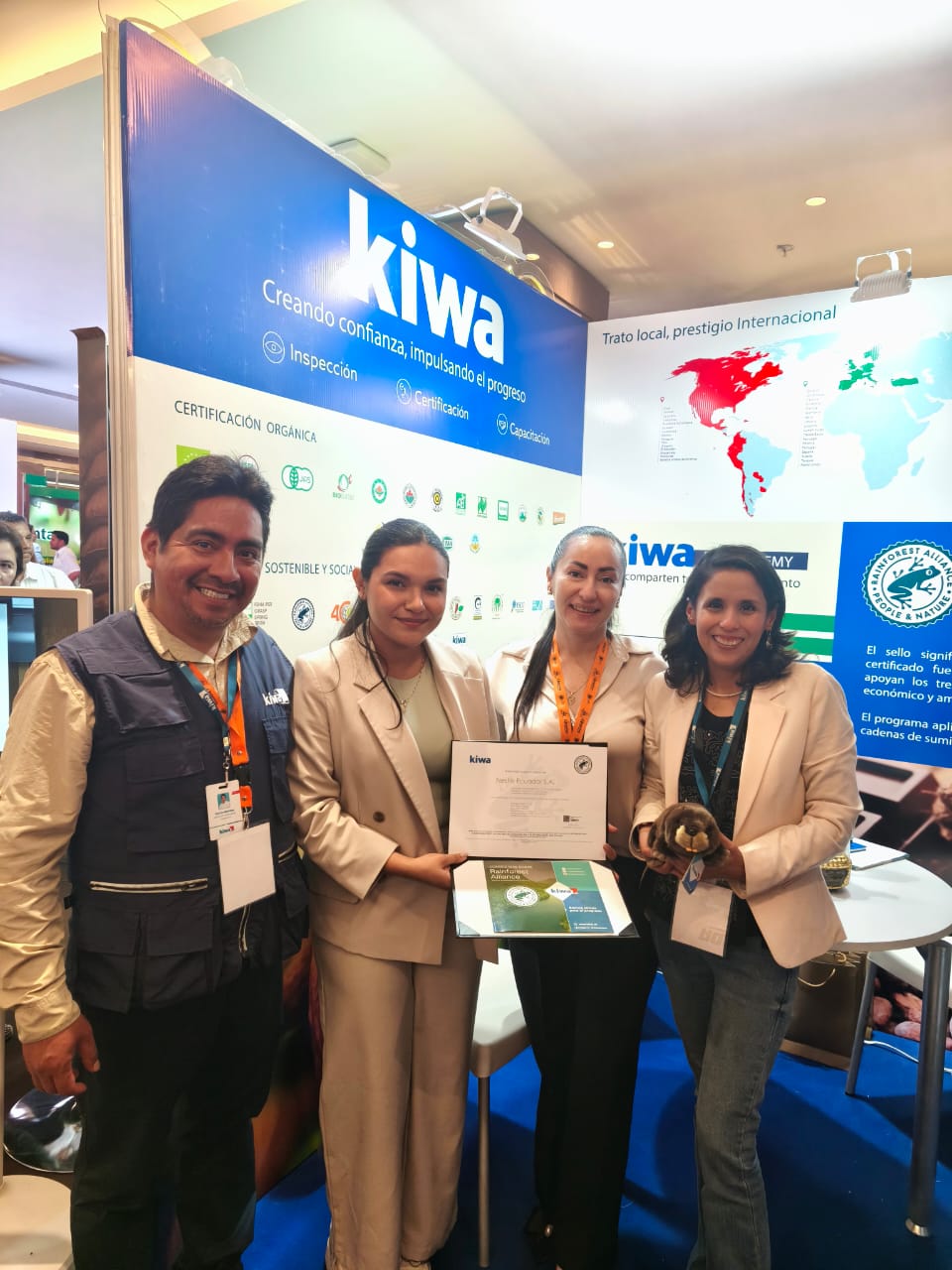 Group of four people at a Kiwa certification event booth, smiling and holding a certificate, with promotional materials and a world map in the background
