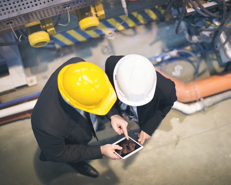 Två personer med bygghjälp är fotograferad uppifrån i en industrilokal. 