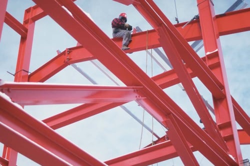 Byggnadsarbetare med skyddsutrustning sitter på en stålbalk i röd byggnadsställning mot blå himmel.