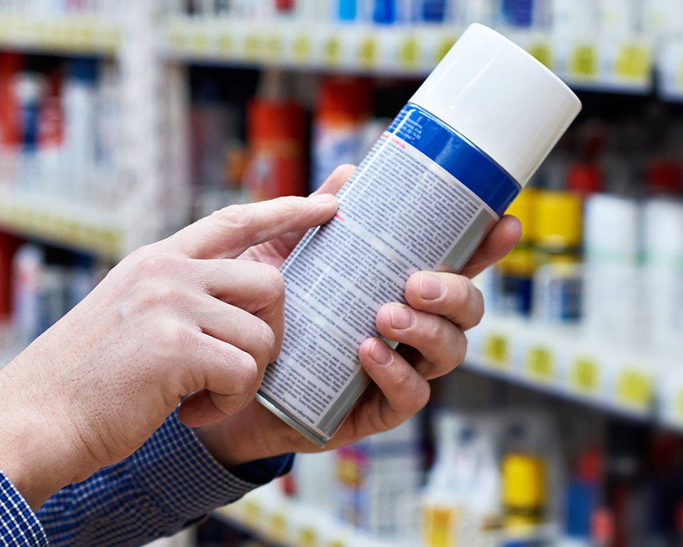 Närbild på händer när en person håller i en sprayflaska och för ett finger längst den finstilta texten på flaskans baksida för att lättare kunna läsa vad som står. 