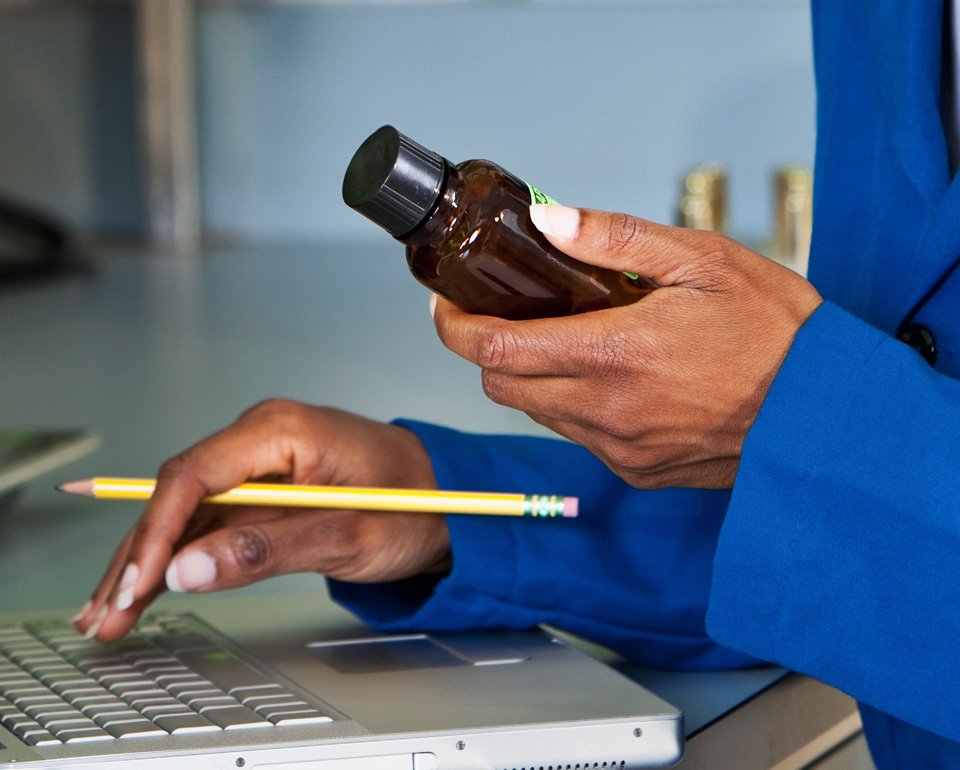 Händer syns där en kvinna håller i en liten flaska med ena handen, medan den andra handen skriver något på en laptop medan den håller i en blyertspenna mellan fingrarna. 