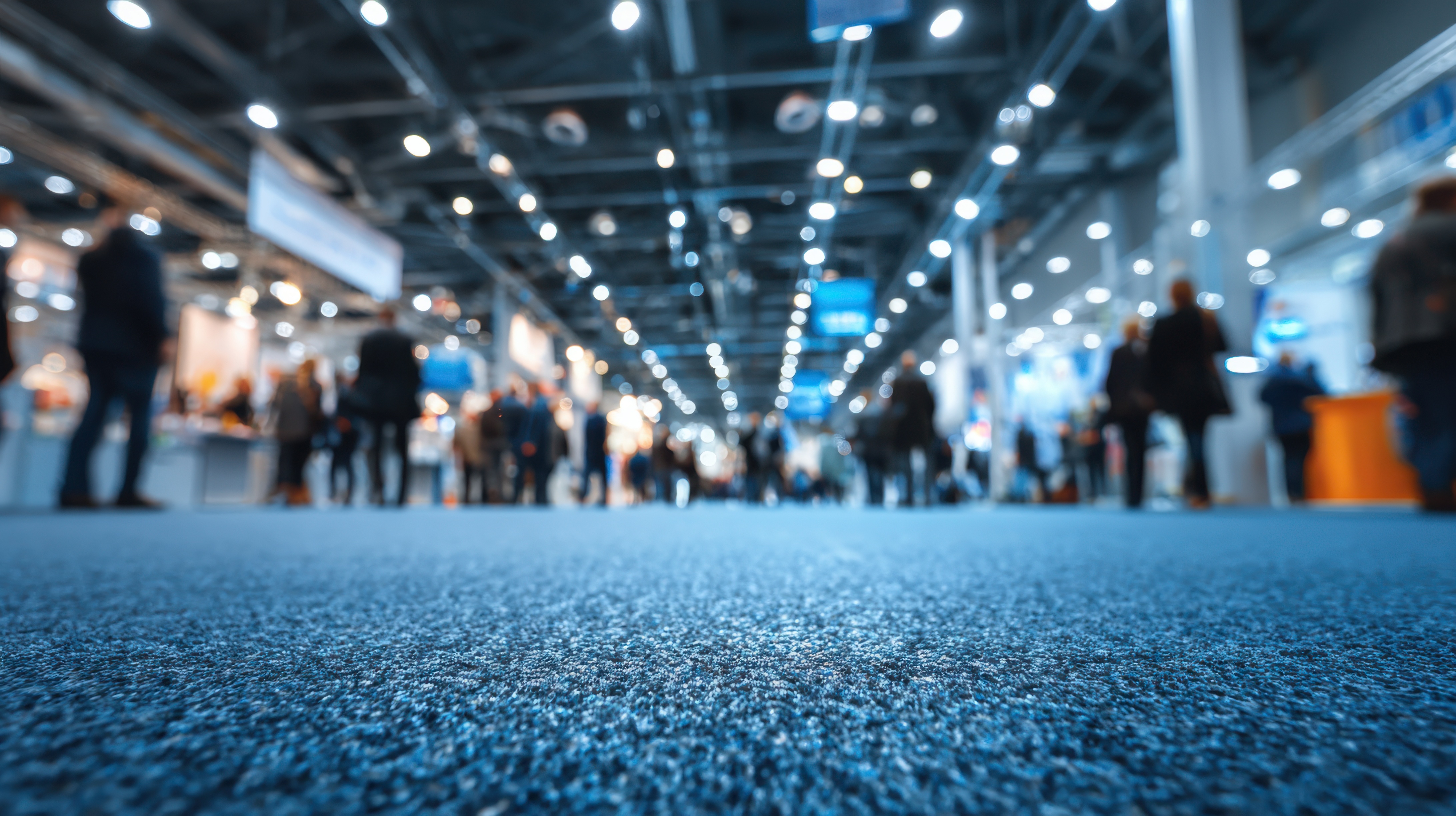 Blurred image of people walking in a convention center with bright overhead lights and a focus on the textured carpet