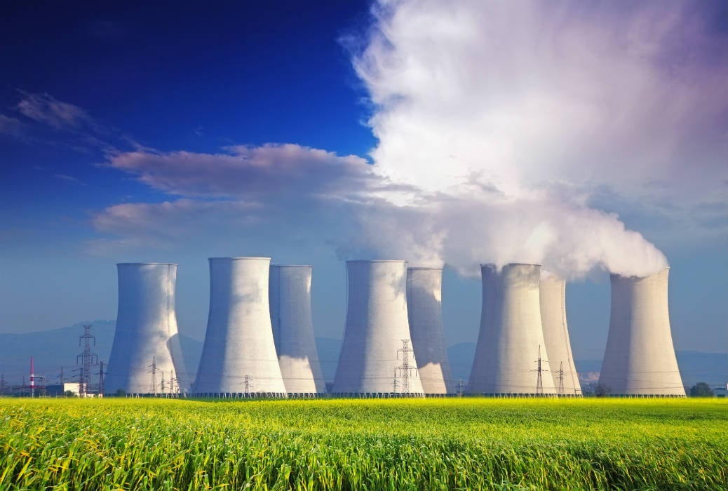 Nuclear power plant cooling towers emitting steam, set against a blue sky with clouds, surrounded by green fields