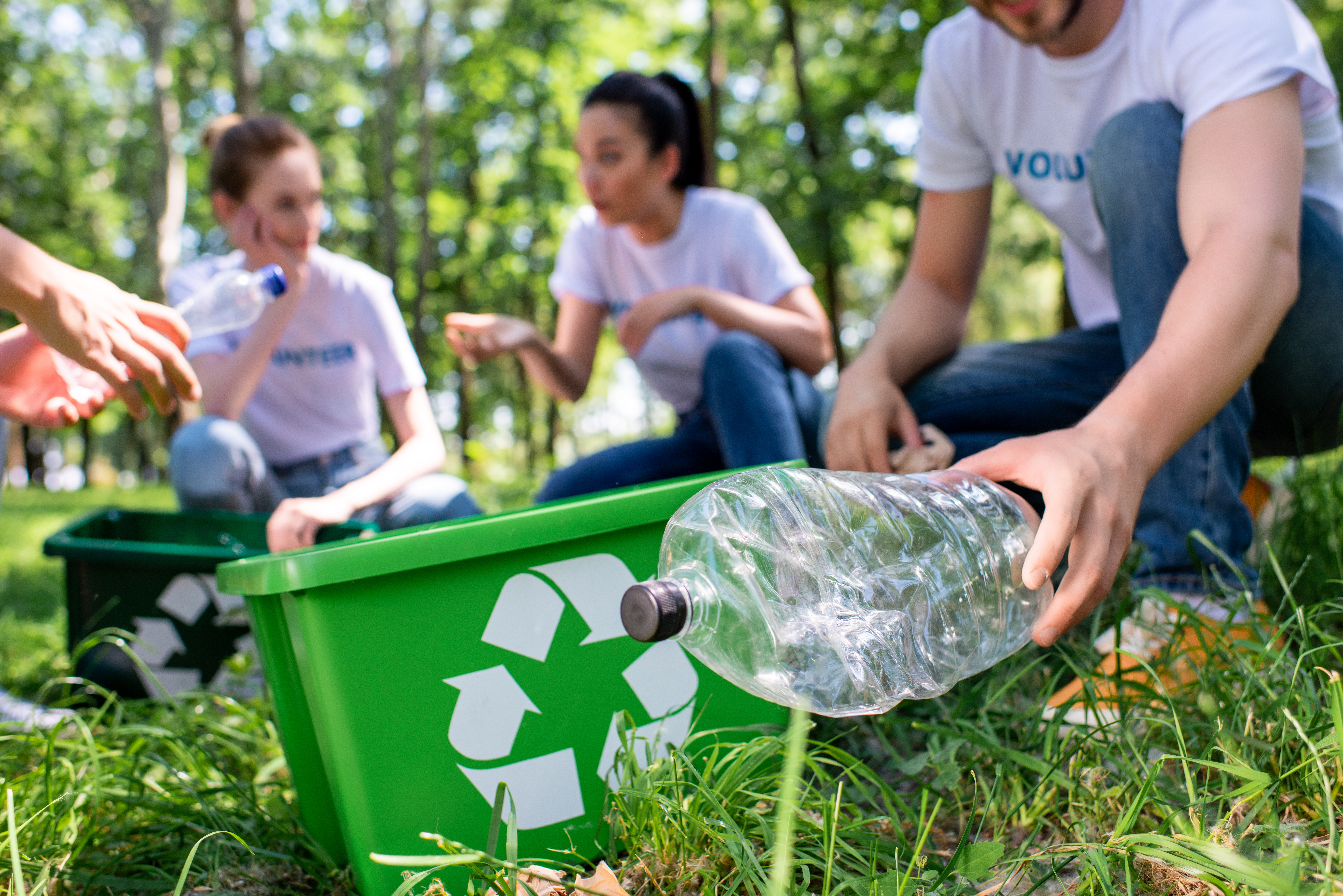 Volunteers collecting plastic bottles for recycling in a park, placing them into green bins with recycling symbols