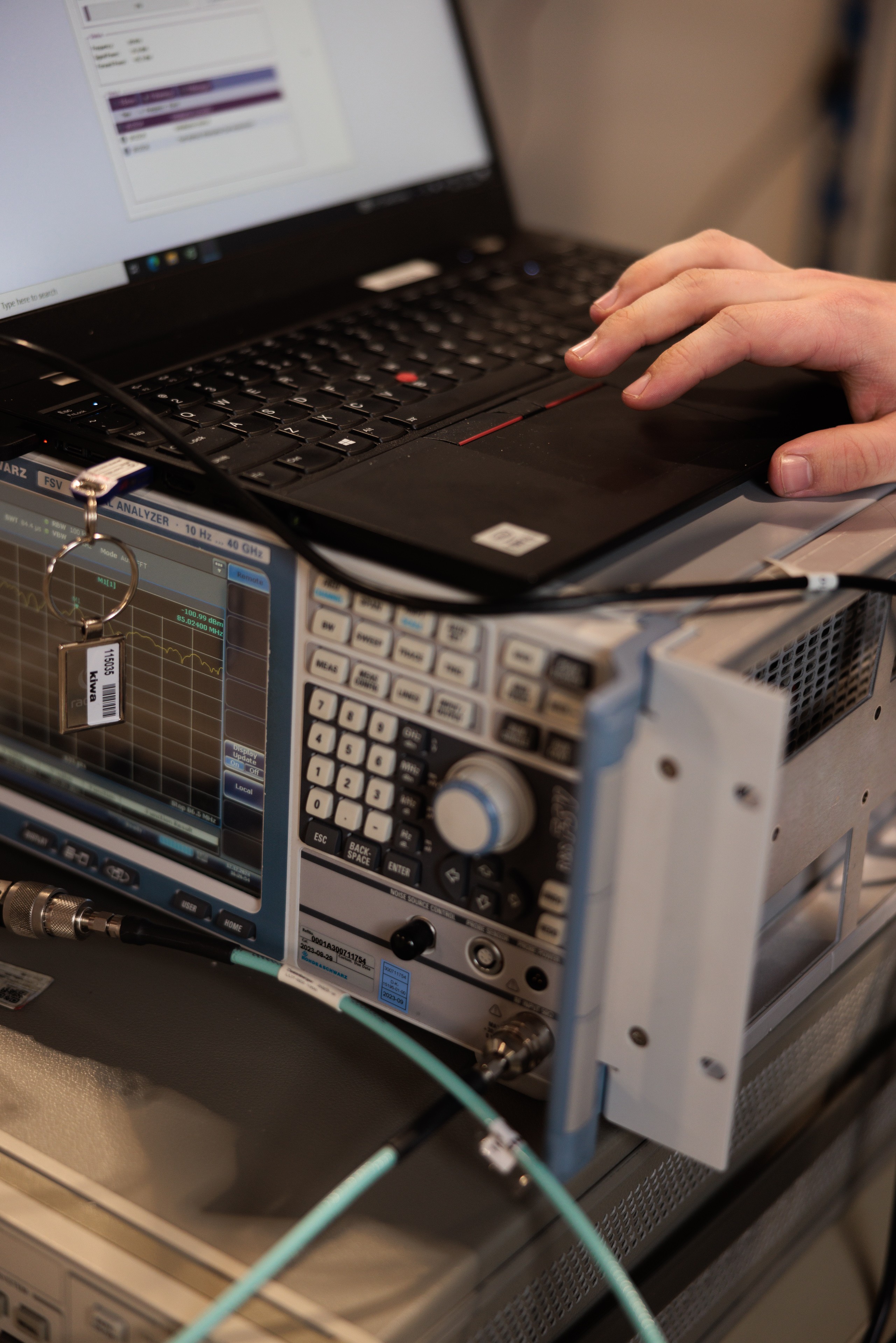 A hand operating a laptop connected to an electronic signal analyzer in a laboratory setting