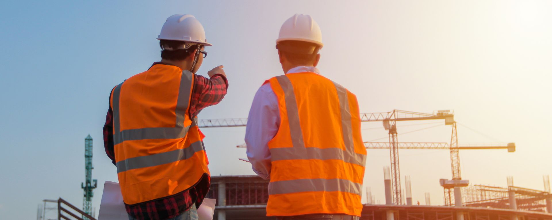 Two Kiwa auditors in orange safety vests and helmets surveying a building site with cranes in the background under a clear sky