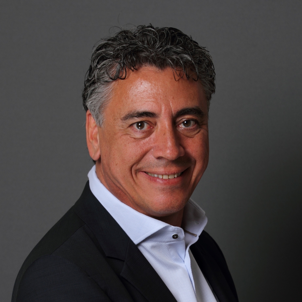 A smiling man with curly hair, wearing a dark suit and light shirt, poses against a gray background.