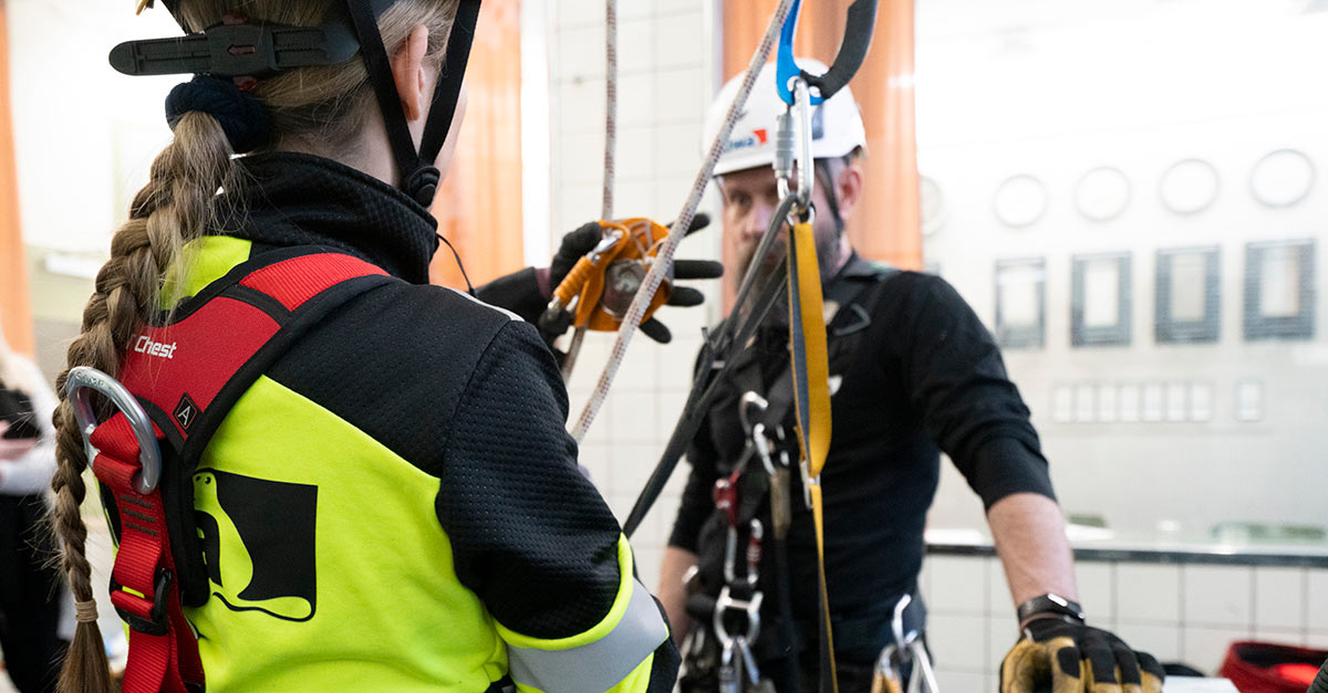 Två personer i skyddsutrustning tränar på arbetssäkerhet med rep och selar inomhus.