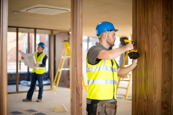 Construction worker using a power drill on a wooden wall, wearing a blue hard hat and safety vest, with another worker holding plans in the background near ladders in a partially constructed room