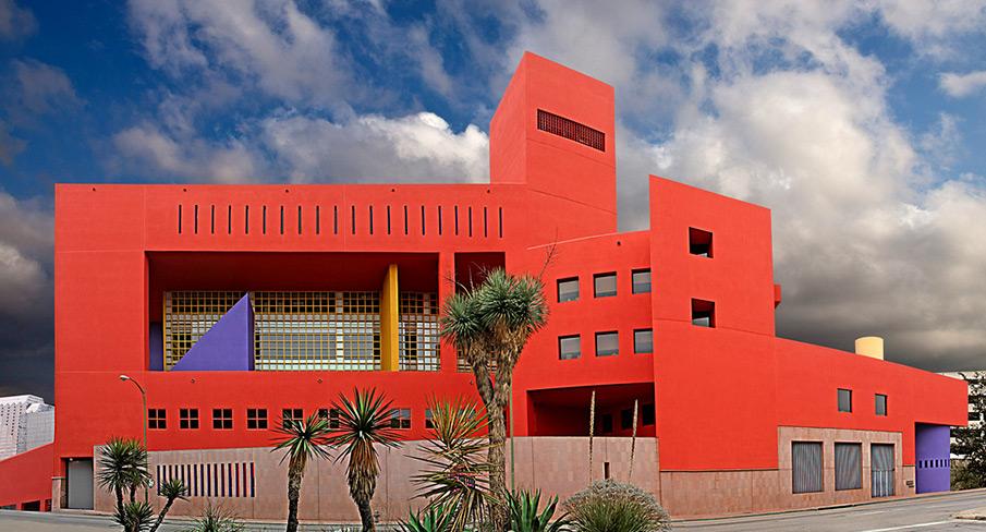 Vibrant red modern building with geometric shapes under a cloudy sky, featuring palm trees in the foreground