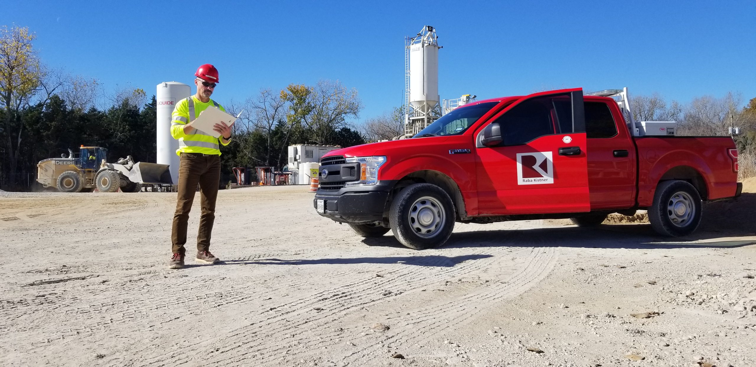 Construction site with a Kiwa Raba Kistner auditors in safety gear holding a clipboard next to a red pickup truck marked with a company logo, surrounded by machinery and storage tanks