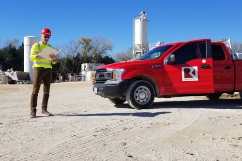 Construction site with a Kiwa Raba Kistner auditors in safety gear holding a clipboard next to a red pickup truck marked with a company logo, surrounded by machinery and storage tanks