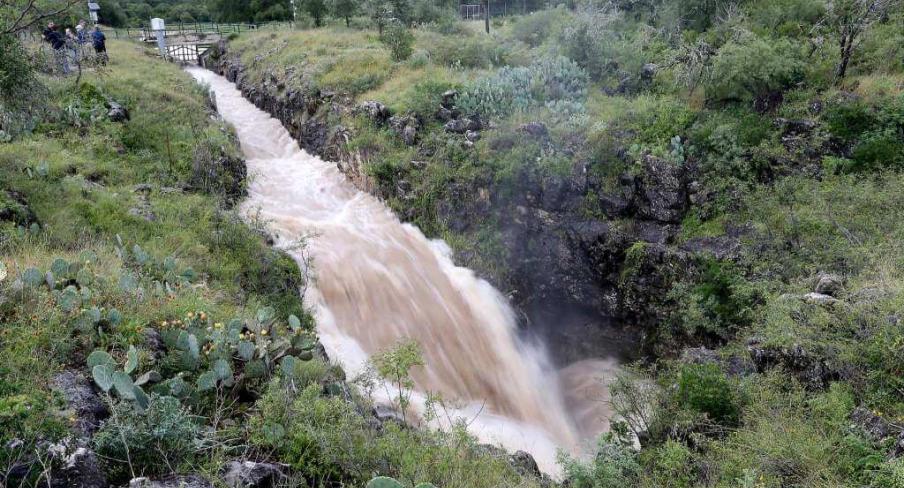 Narrow river flowing through rocky terrain amidst lush greenery, with a small bridge visible in the background