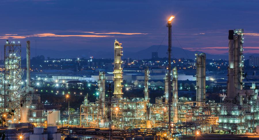 Illuminated oil refinery at dusk with industrial towers and a backdrop of a glowing cityscape