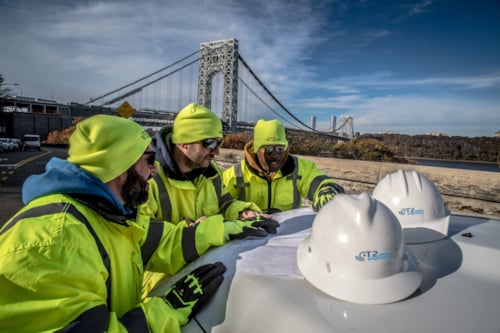 Kiwa T2UE engineers in high-visibility jackets and helmets reviewing plans near a bridge on a clear day